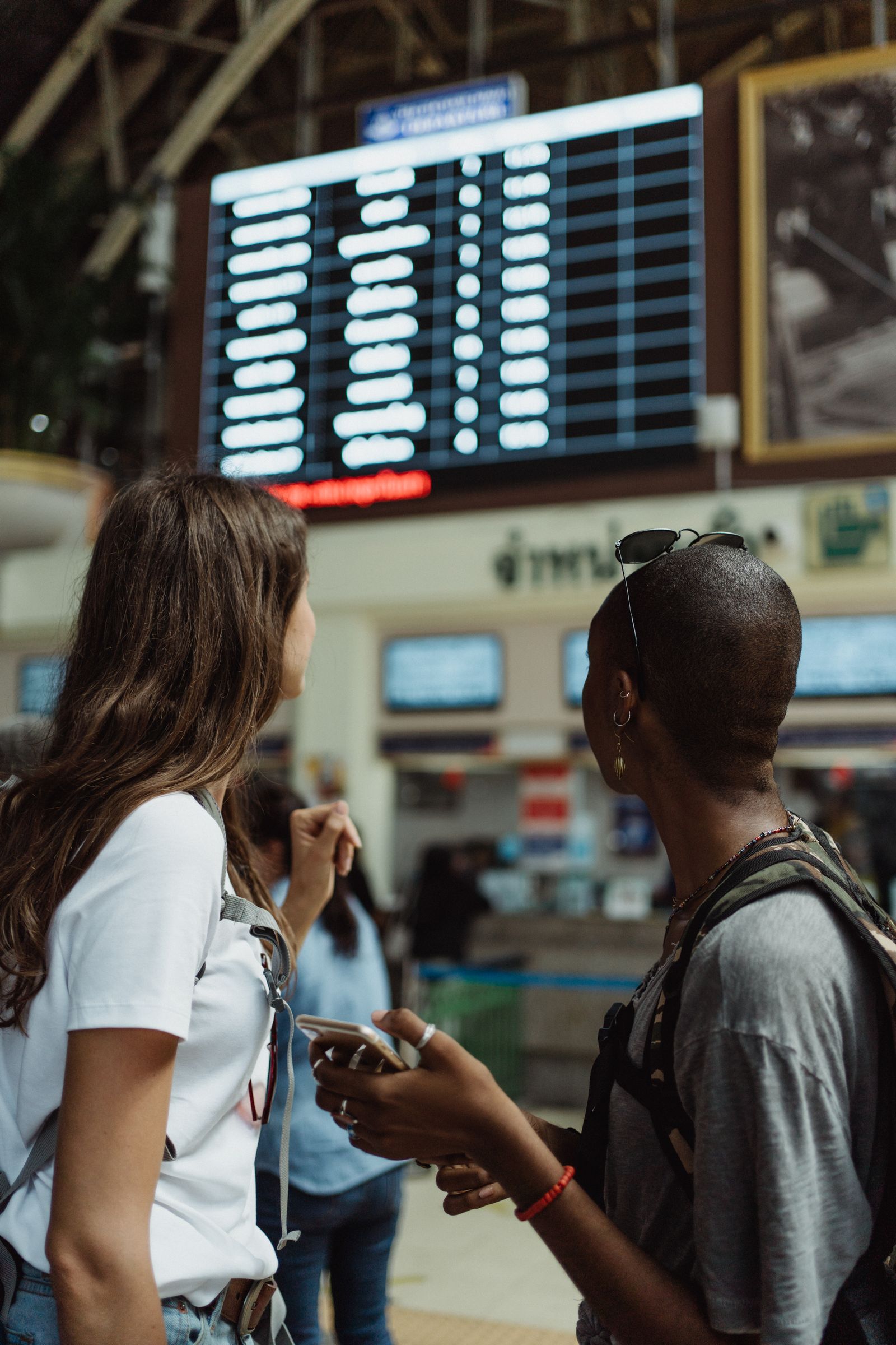 Travellers standing below a digital airport flight information board